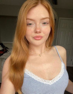 Redheaded woman with fair skin and freckles wearing a blue striped tank top with lace trim, taking a selfie indoors.