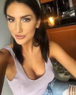 Brunette woman in a low-cut lavender top and ripped jeans takes a selfie in a warmly lit kitchen setting.