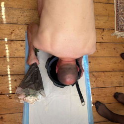 A shirtless man is bent over on a mat with his head inside a bucket, holding a bag of flowers; another person's feet in