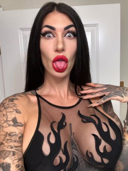 Brunette woman with full tattoos on arms and torso wearing a black sheer top with flame cutouts, making a playful face