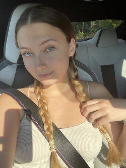 A young woman with long braided hair sits in a car, wearing a white tank top and seatbelt, looking at the camera.