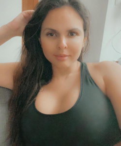 Curvy woman with long dark hair wearing a black tank top, posing indoors with one hand in her hair.