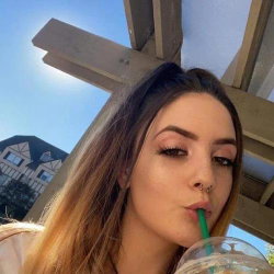 Brunette woman with long hair and a nose ring sipping from a drink under a wooden pergola on a sunny day.