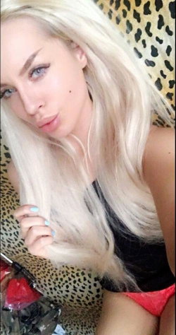 Blonde woman with long hair in a black top and red underwear, posing on a leopard-print surface in a selfie-style shot.