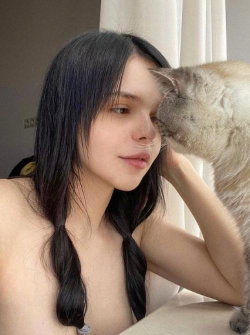 A young woman with long dark hair in braids is topless, gently holding a cat that nuzzles her face indoors.