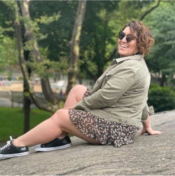 Curvy woman with curly hair wearing a green jacket and floral skirt, sitting on a stone ledge in a park with trees