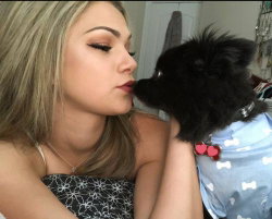 Blonde woman with defined makeup kisses a small black dog wearing a blue outfit indoors.