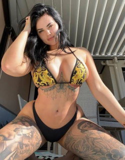 Brunette woman with curvy build and full tattoos on thighs, wearing a yellow and black patterned bikini top and black