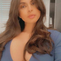 Brunette woman with long wavy hair in a blue top, taking a close-up selfie indoors.