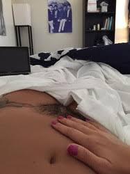 Close-up of a person's lower abdomen with hand resting on stomach, partially covered by white sheets in a bedroom