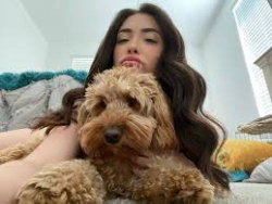 Brunette woman with long wavy hair lying on a bed, hugging a fluffy brown dog, partially covered by blankets.