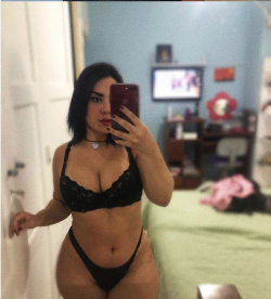Curvy woman in black lace lingerie takes mirror selfie in bedroom, holding phone with one hand, standing near bed and