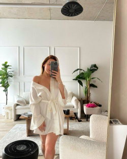 A woman with shoulder-length hair takes a mirror selfie in a white off-the-shoulder robe, standing in a bright, modern