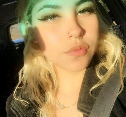 Blonde woman with curly hair and makeup, wearing a dark jacket and seatbelt, taking a selfie in a car.