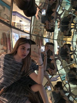 A young woman with long blonde hair takes a selfie in a mirrored room, wearing a plaid dress and holding a phone.