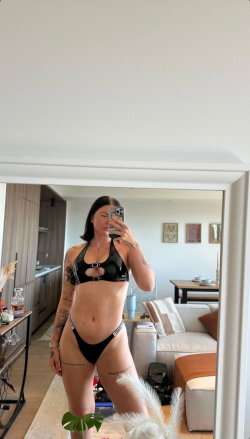 Curvy woman with tattoos takes mirror selfie in black lingerie, standing in a modern living room.