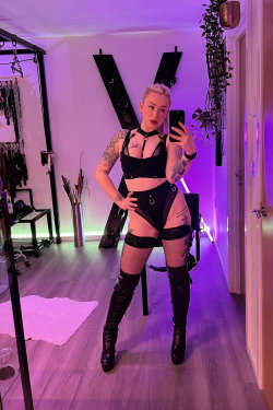 Blonde woman with tattoos takes a mirror selfie in a black harness, fishnet stockings, and thigh-high boots in a room