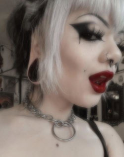 Person with platinum blonde hair, bold black eyeliner, red lipstick, and a chain choker, wearing a black top in an