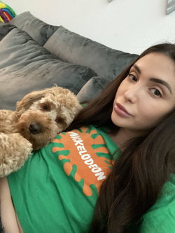 Brunette woman in a green Nickelodeon shirt lying on a gray couch with a small fluffy dog resting on her chest.