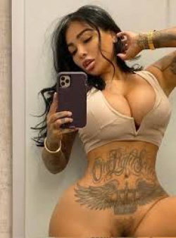 A curvy woman with dark hair takes a mirror selfie in a beige crop top, showing tattoos on her arms and torso.