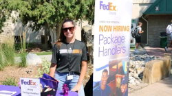 A woman in a FedEx Ground uniform and sunglasses stands outdoors near a hiring sign, holding promotional materials.