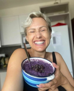 A smiling woman with short silver hair holds a bowl of purple smoothie in a kitchen, wearing a black tank top.