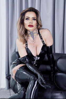A woman with wavy hair and bold makeup wears a black latex outfit with long gloves, sitting on a leather chair against