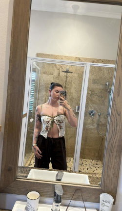 A woman with tattoos takes a mirror selfie in a bathroom, wearing a floral crop top and dark pants, standing near a