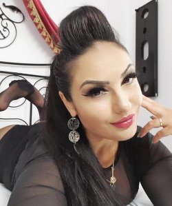 Brunette woman with long dark hair in a high ponytail, wearing sheer black top and black stockings, posing on a bed