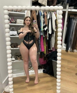 Curvy woman in black lace-up bodysuit takes mirror selfie in walk-in closet, barefoot with long wavy hair.