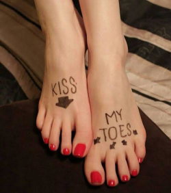 Close-up of bare feet with red toenails and 'KISS MY TOES' written in black ink, resting on a dark surface near bedding.
