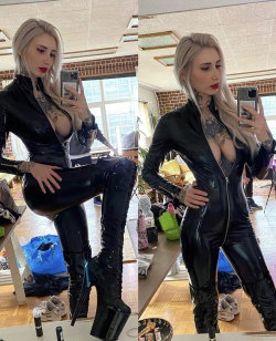 Blonde woman in a black latex catsuit and high platform boots poses in a mirror selfie, showcasing tattoos on her chest