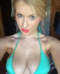 Blonde woman with blue eyes in a teal halter top, taking a close-up selfie indoors.