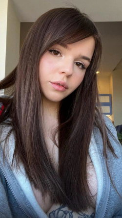 Brunette woman with long hair in a gray zip-up hoodie, taking a close-up selfie indoors.