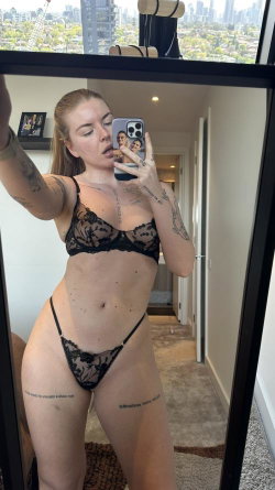 Blonde woman with tattoos takes mirror selfie in black lace lingerie, standing in a modern apartment with city view.