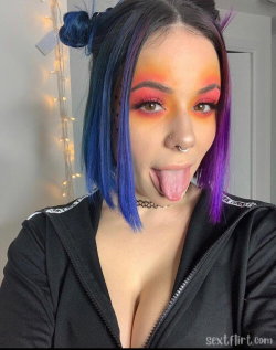 Woman with blue and purple hair, bold makeup, sticking tongue out, wearing a black zip-up top in a room with string