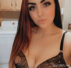 A woman with long auburn hair and defined makeup wears a black lace top, taking a selfie in a laundry room.