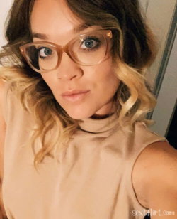 Woman with wavy brown hair and glasses wears a beige sleeveless top, taking a close-up selfie indoors.