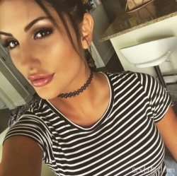 A woman with dark hair and makeup takes a selfie in a striped shirt, wearing a choker, in what appears to be a kitchen