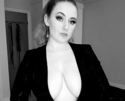 A woman with blonde hair in a high ponytail wears a black blazer open to reveal her bare chest, posing indoors with