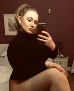 Curvy woman in a black turtleneck takes a mirror selfie on a bed, hair styled in an updo with makeup and red nails.