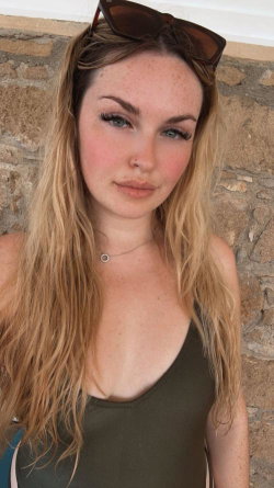 Blonde woman with freckles and long hair wearing a green tank top, sunglasses on head, standing against a stone wall.