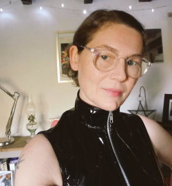 A woman with short brown hair and glasses wears a shiny black sleeveless top, smiling softly in a cozy room with string
