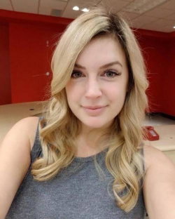 Blonde woman with wavy hair wearing a gray tank top, smiling in a selfie taken indoors against a red wall.
