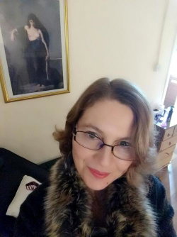 A woman with shoulder-length brown hair and glasses smiles at the camera, wearing a dark coat with a fur collar in a