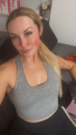 Blonde woman with ponytail in a gray crop top and black pants, taking a selfie on a couch with visible arm tattoo.
