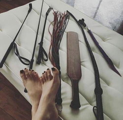 A person's bare feet with dark nail polish rest on a white tufted surface beside various BDSM implements including