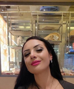 Brunette woman with bold makeup and pink lipstick smiles softly in a selfie taken indoors near a display case.