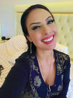 A smiling woman with dark hair and bold makeup takes a selfie in a bedroom, wearing a navy blue patterned top with a