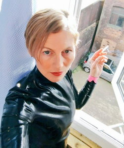 Woman with short blonde hair in a black latex top, holding a cigarette near a window, looking at the camera.